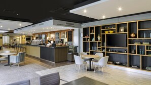 Un hall d’hôtel moderne avec un comptoir Starbucks, des espaces de détente et un téléviseur posé sur une étagère en bois.