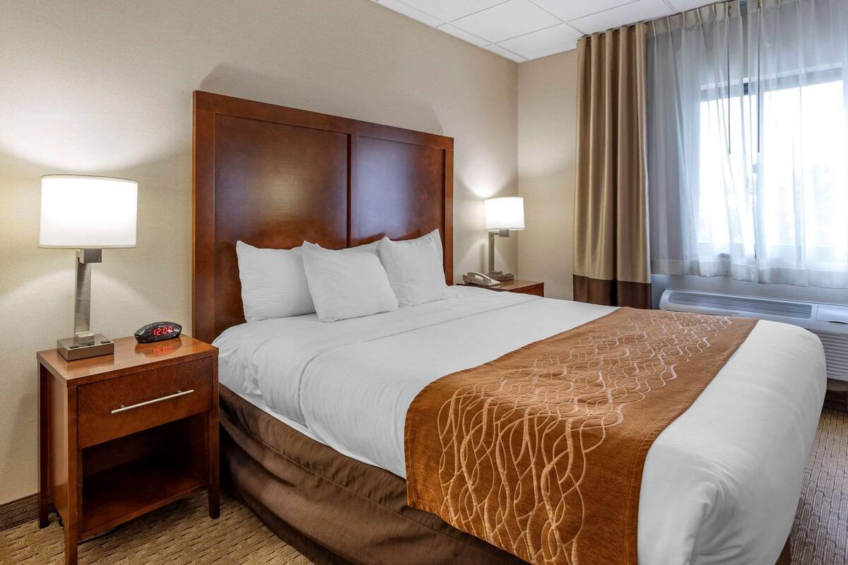 A hotel room with a large bed, wooden headboard, two bedside tables with lamps, and a window with curtains.