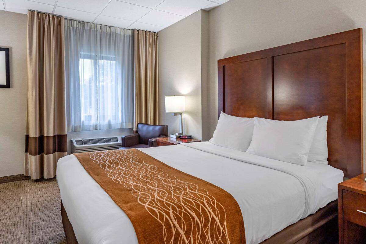 A hotel room with a large bed, a wooden headboard, a window with curtains, a desk with a lamp, and an air conditioning unit.