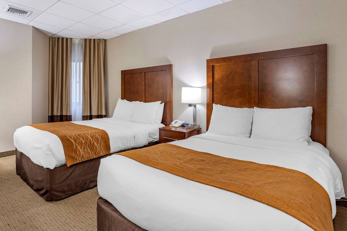 A hotel room with two beds, a nightstand, a lamp, and a window with curtains.