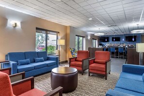 A hotel lobby with a blue sofa, red armchairs, a coffee table, a bar area with a variety of bottles, and a large window providing a view of the outdoors. - Comfort Inn Syosset - Long Island (Syosset)
