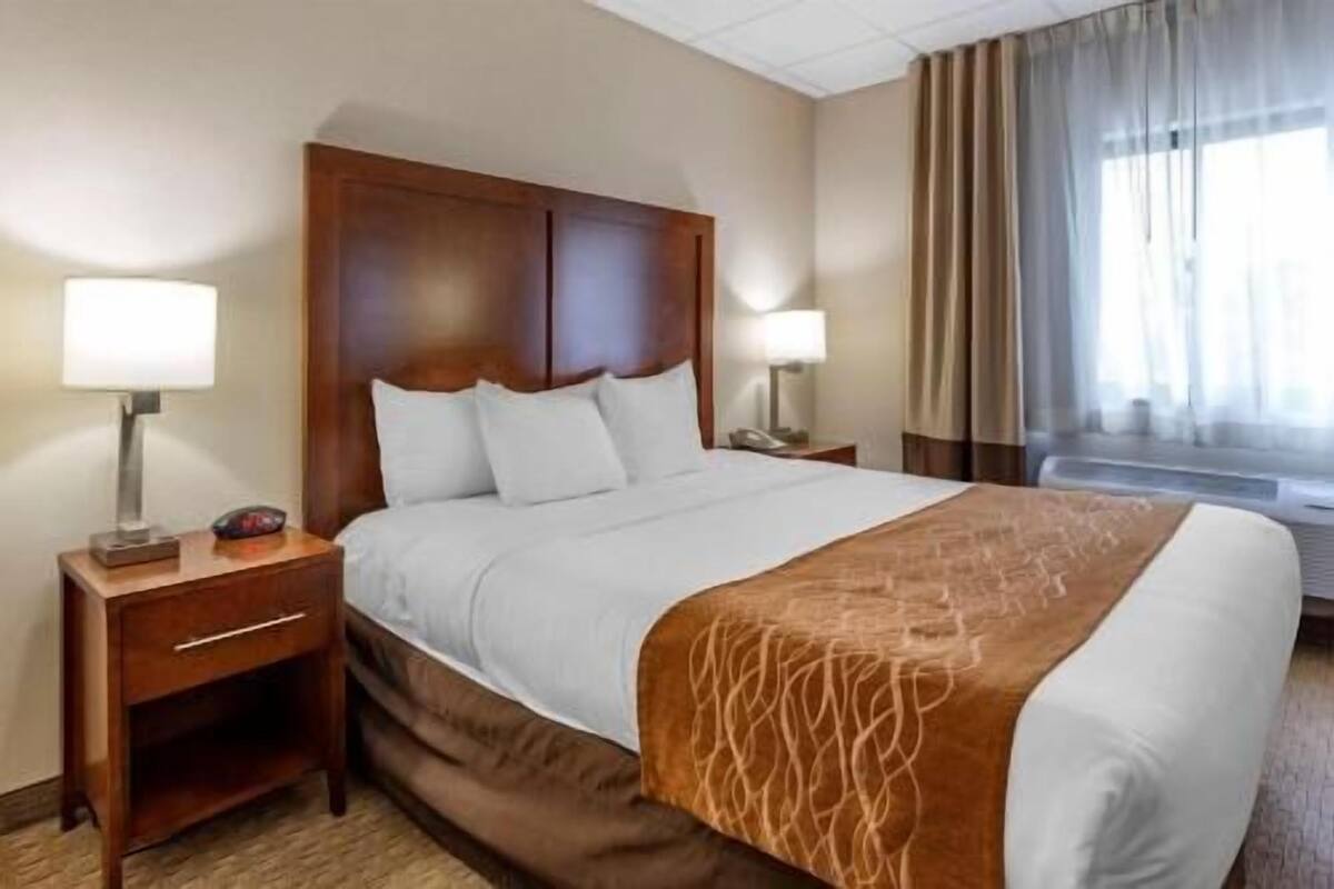 A hotel room with a large bed, wooden headboard, two bedside tables with lamps, a nightstand, and a window with curtains.