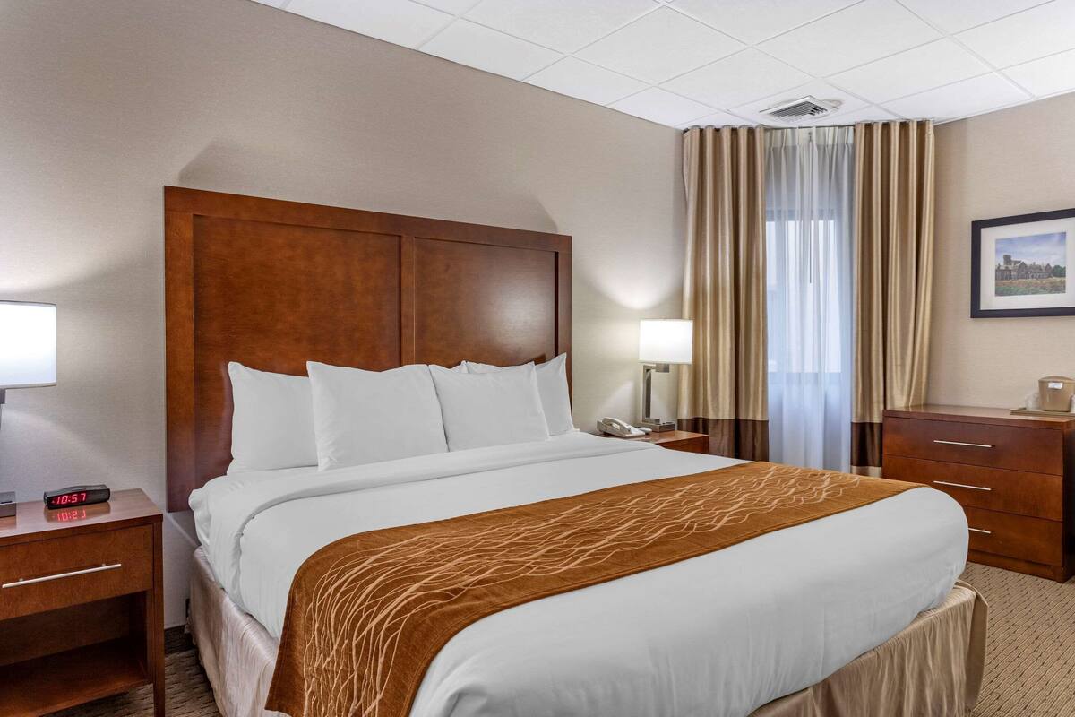 A hotel room with a large bed, wooden headboard, two bedside tables with lamps, a nightstand, and a dresser.