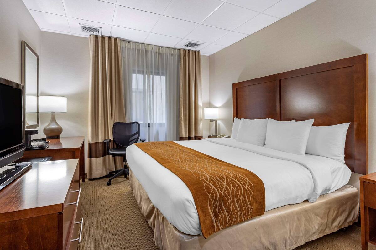 A hotel room with a large bed, a desk with a computer, a chair, a TV, and a window with curtains.