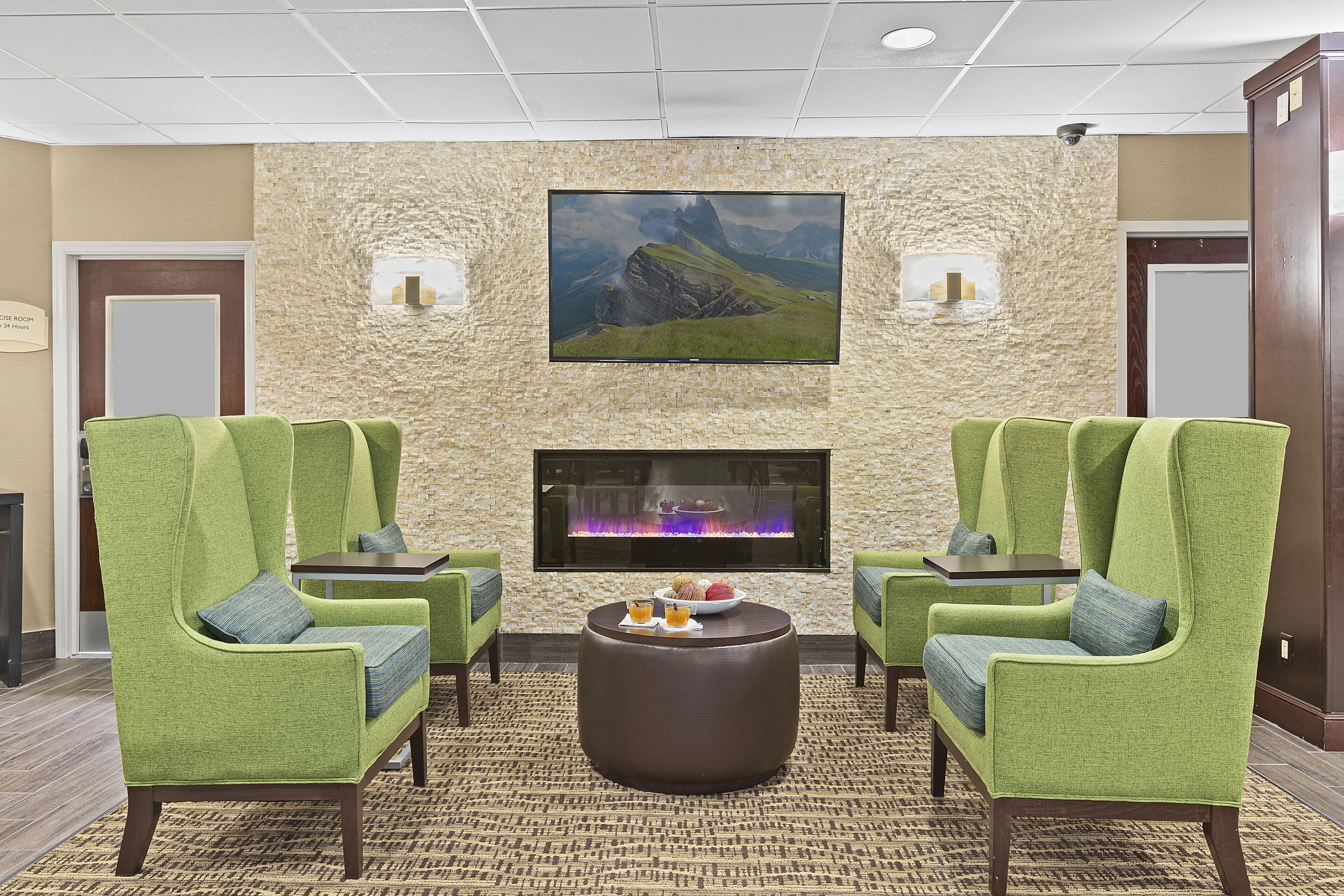 A lobby with green upholstered chairs, a round coffee table, and a large flat-screen TV mounted on a textured stone wall.