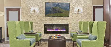 A lobby with green upholstered chairs, a round coffee table, and a large flat-screen TV mounted on a textured stone wall.