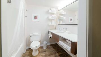 Ein modernes Badezimmer mit einer weißen Toilette, einem Waschbecken und einem Spiegel. Handtücher liegen auf einem Regal und ein Mülleimer steht auf dem Boden.