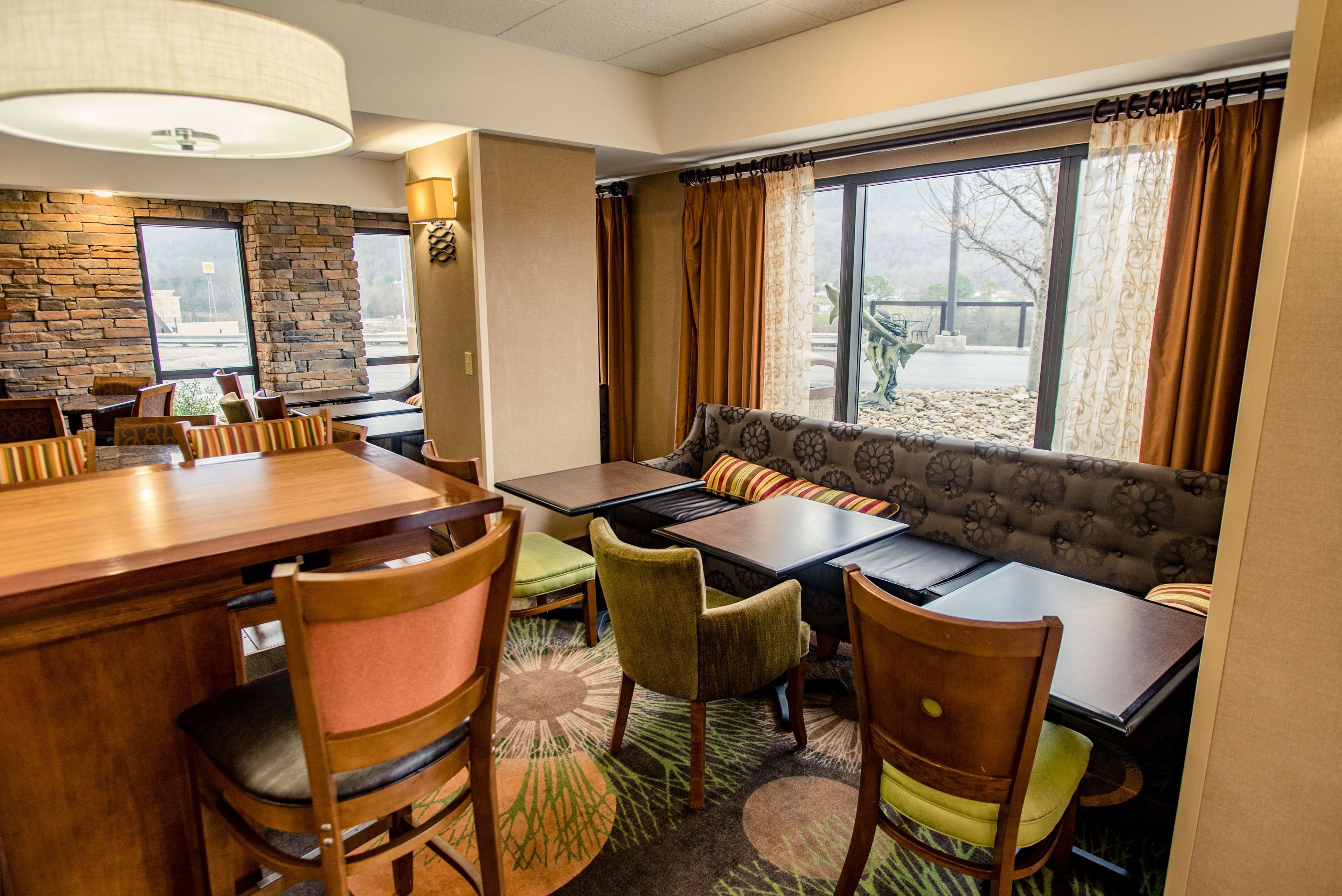 A dining area with a wooden table, chairs, and a sofa. There are windows with curtains, a stone wall, and a carpeted floor.