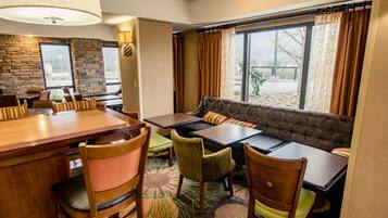 A dining area with a wooden table, chairs, and a sofa. There are windows with curtains, a stone wall, and a carpeted floor.