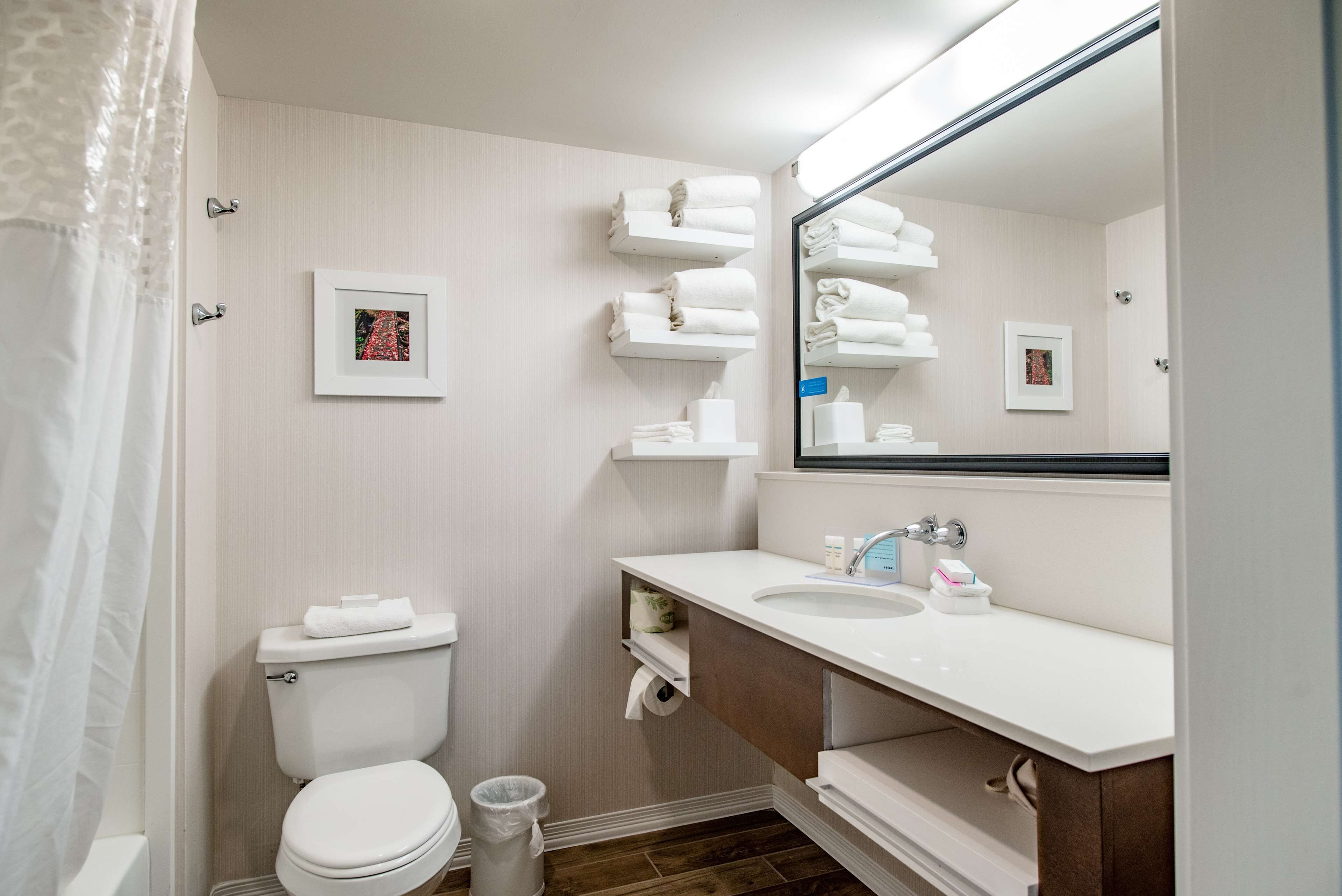 A bathroom with a toilet, sink, and mirror. There are towels on a shelf and a picture on the wall.