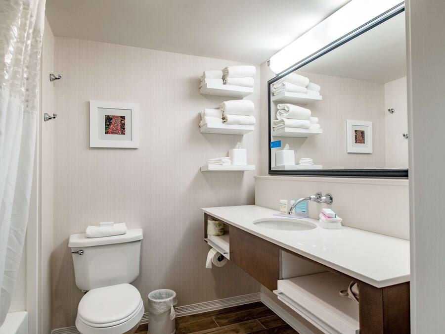A bathroom with a toilet, sink, and mirror. There are towels on a shelf and a picture on the wall.
