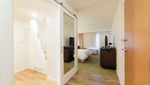 Habitación de hotel con piso de madera, una cama con cubrecama blanco, un escritorio con computadora, un televisor y un espejo.