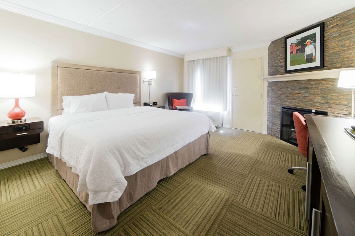 A hotel room with a bed, bedside table, lamp, chair, desk, TV, and a fireplace.