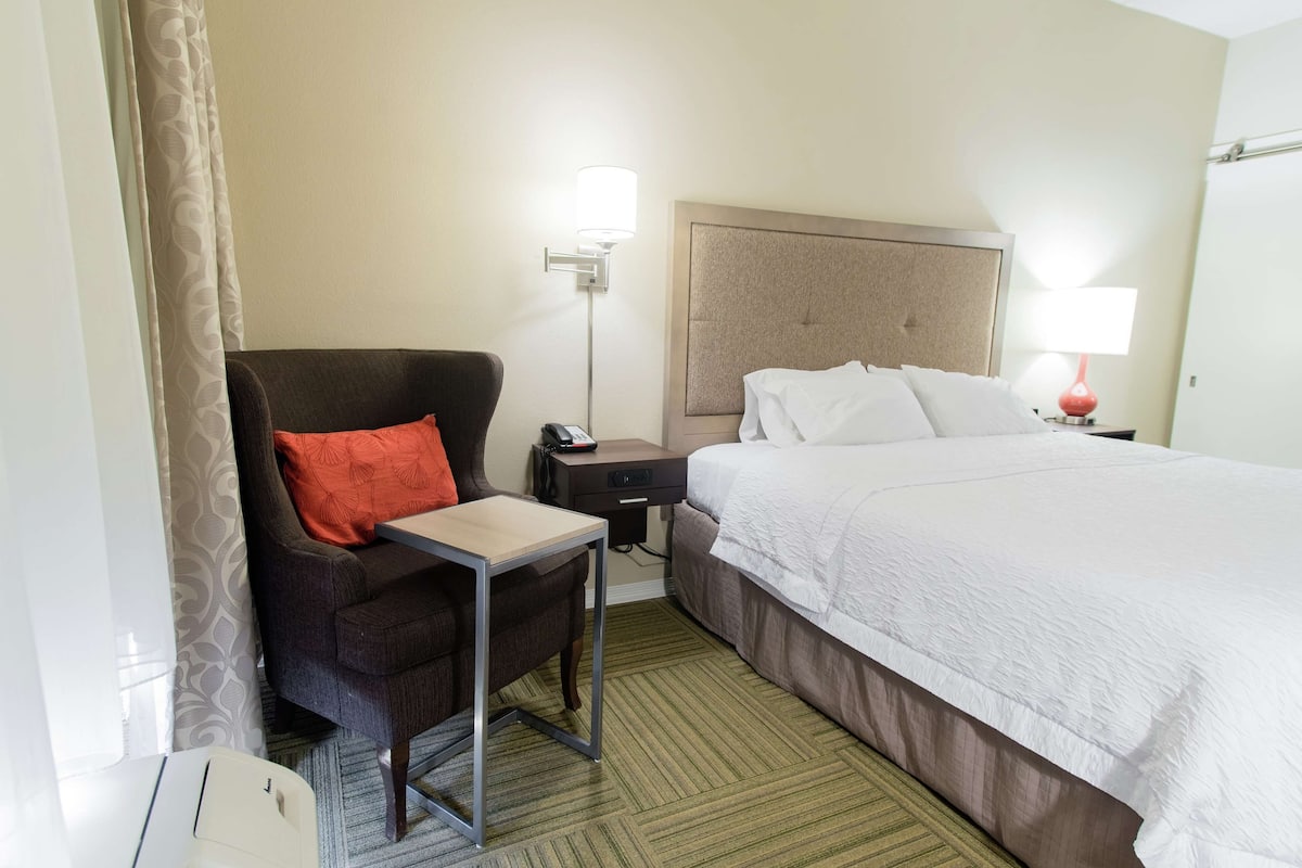 A hotel room with a bed, a desk, a chair, a nightstand, and a lamp.
