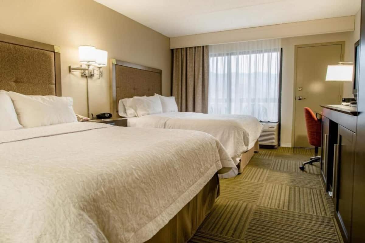 A hotel room with two beds, a desk, a chair, a lamp, and a large window with curtains.