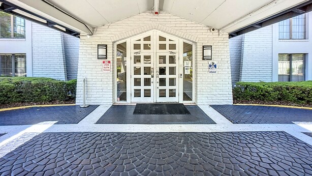 Una entrada de edificio con fachada de ladrillo blanco, puerta de cristal y felpudo negro.