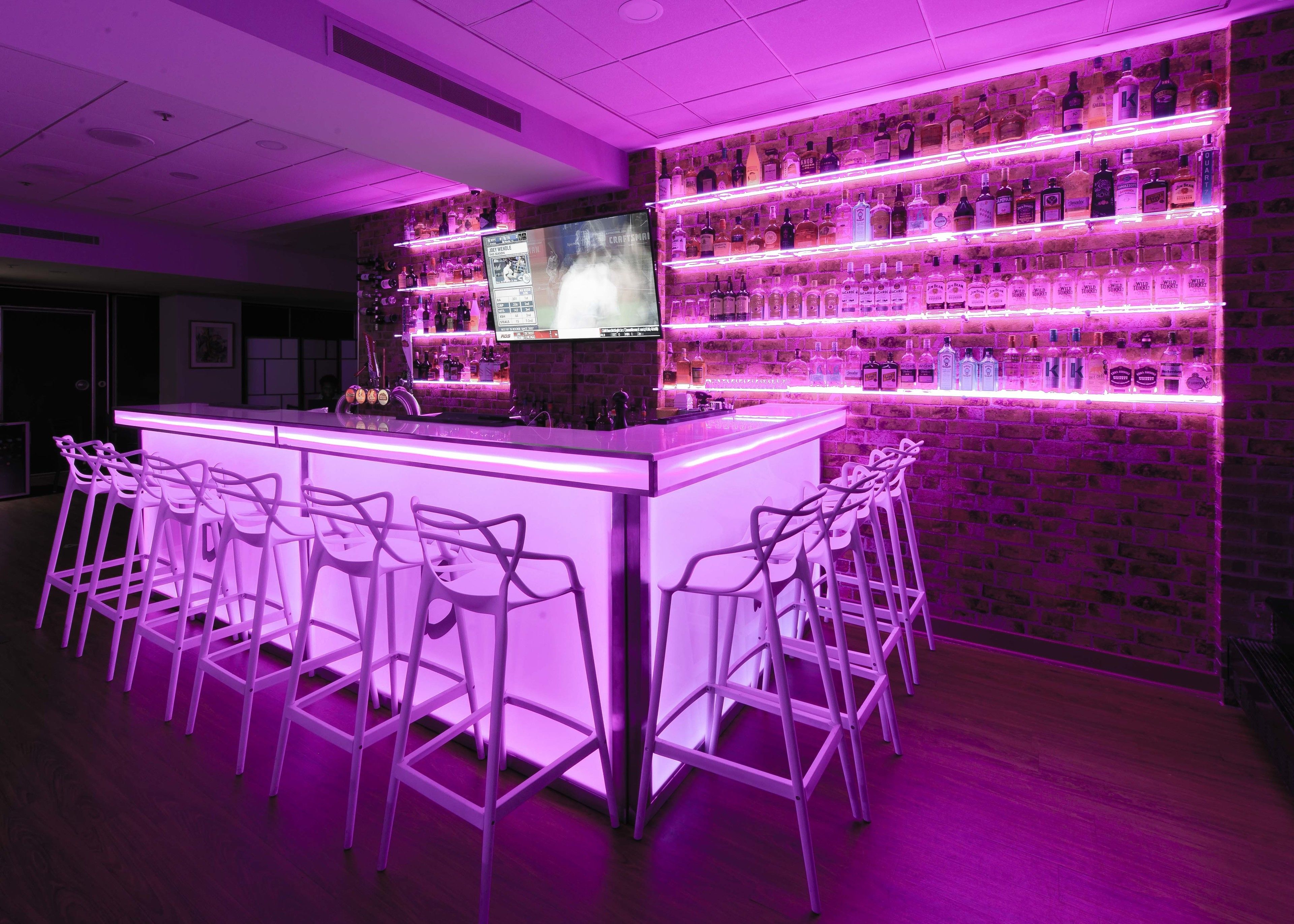 Zona de bar con barra iluminada con neón, varios estantes llenos de botellas de licor y taburetes de bar.