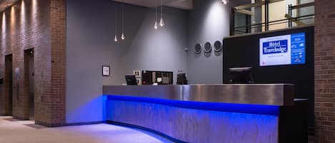 A hotel reception desk with a blue-lit counter, a digital screen displaying hotel information, and a clock on the wall.