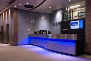 Una recepción de hotel con un mostrador iluminado en azul, una pantalla digital mostrando información del hotel y un reloj en la pared.