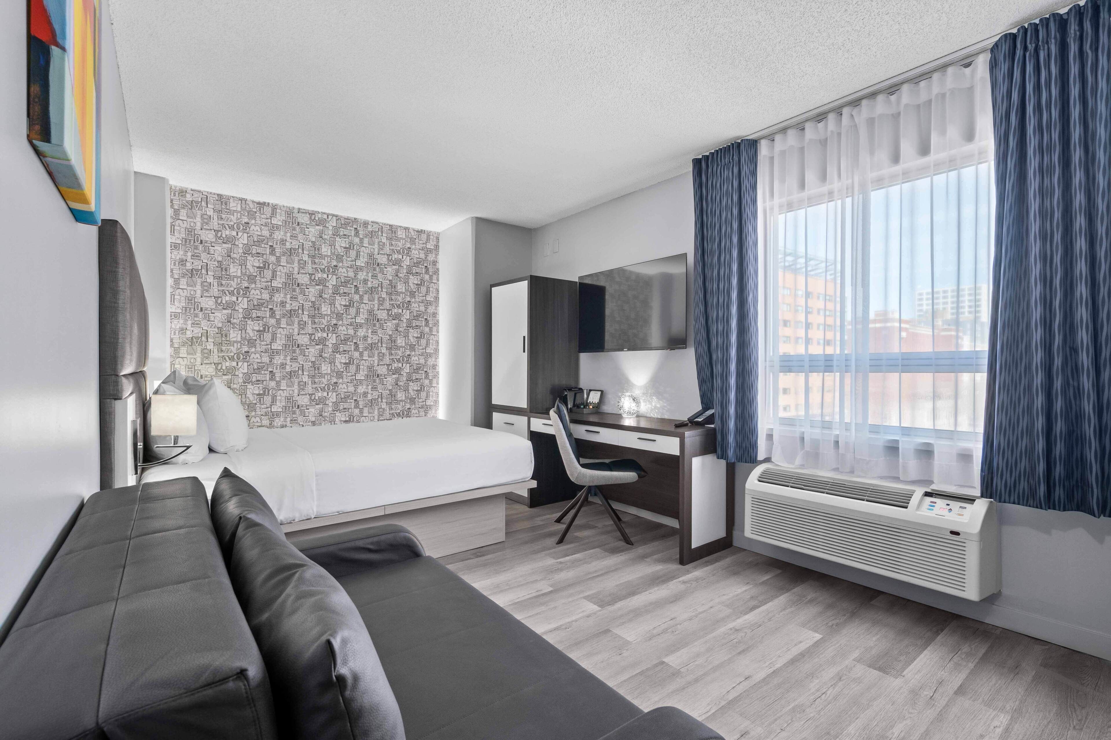 Ein modernes Hotelzimmer mit Bett, Schreibtisch, Stuhl, Sofa und einem großen Fenster mit Vorhängen.
