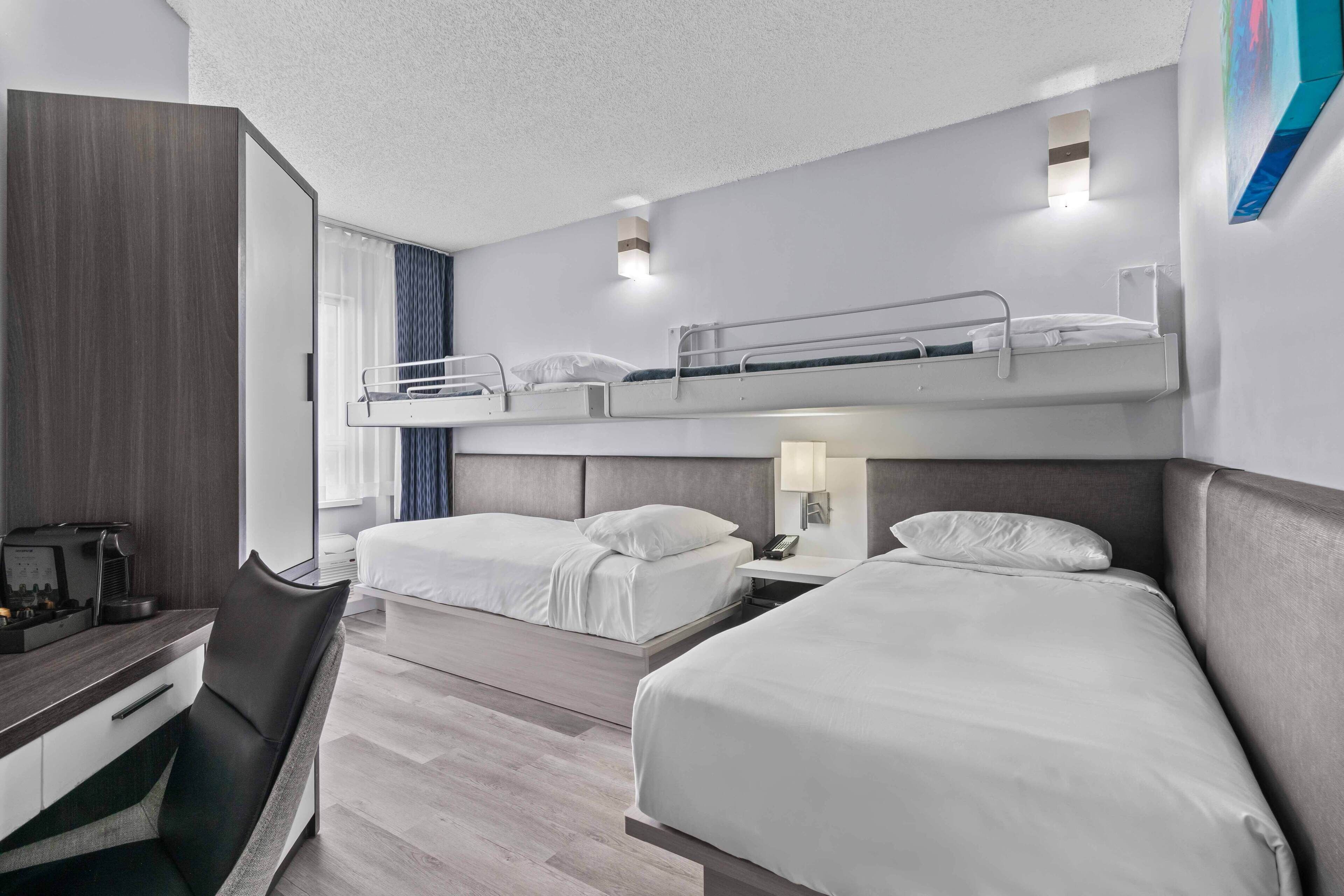 A hotel room with a bunk bed, a desk with a coffee maker, and a bedside table with a lamp.