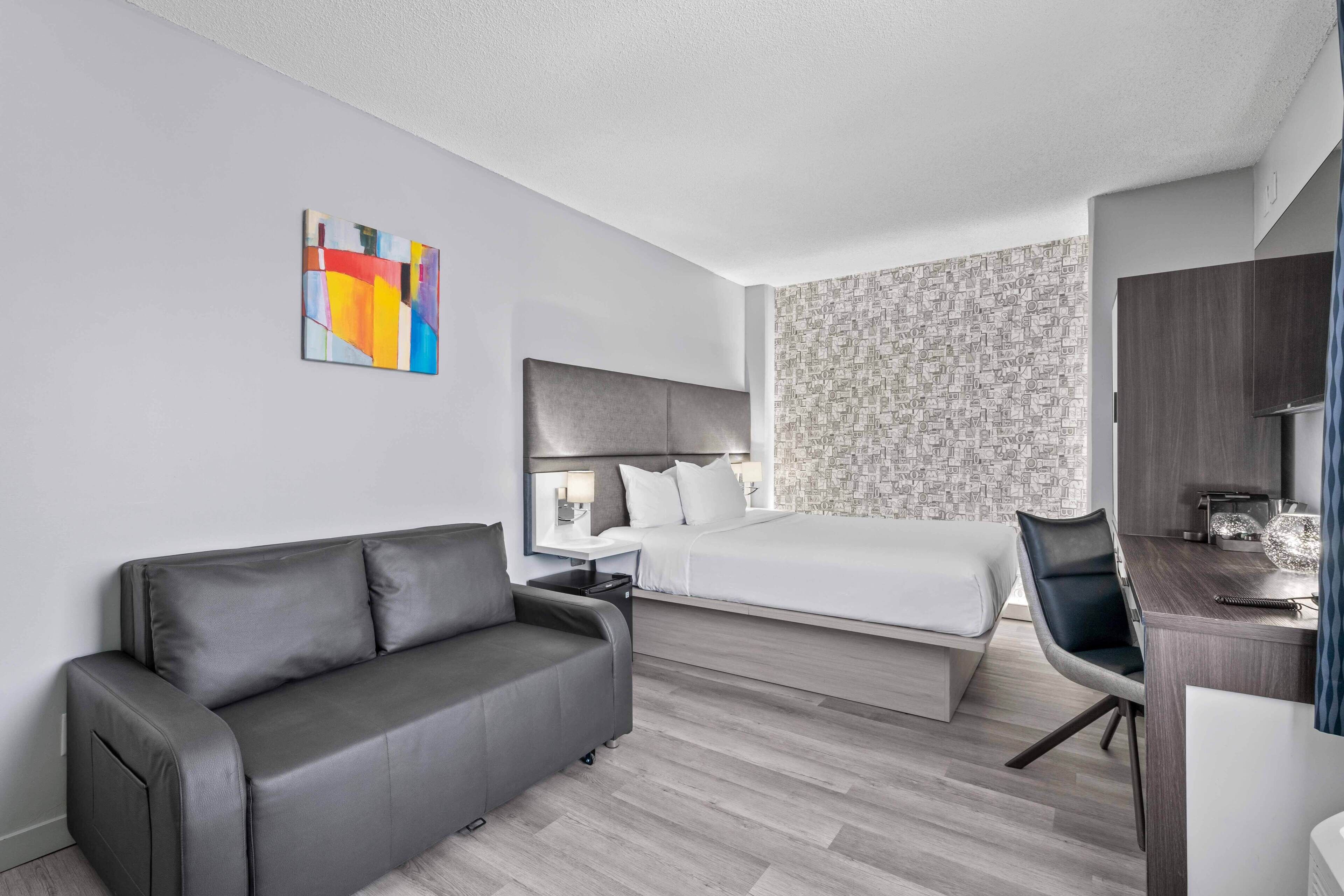 A modern hotel room with a grey sofa, a bed, a desk, and a colorful abstract painting on the wall.