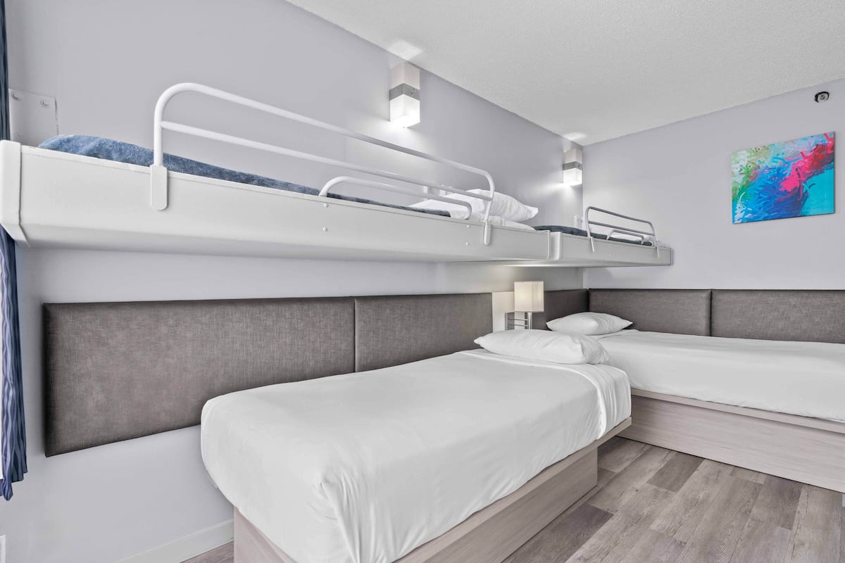 A hotel room with a bunk bed, two single beds, a bedside table with a lamp, and a colorful abstract painting on the wall.