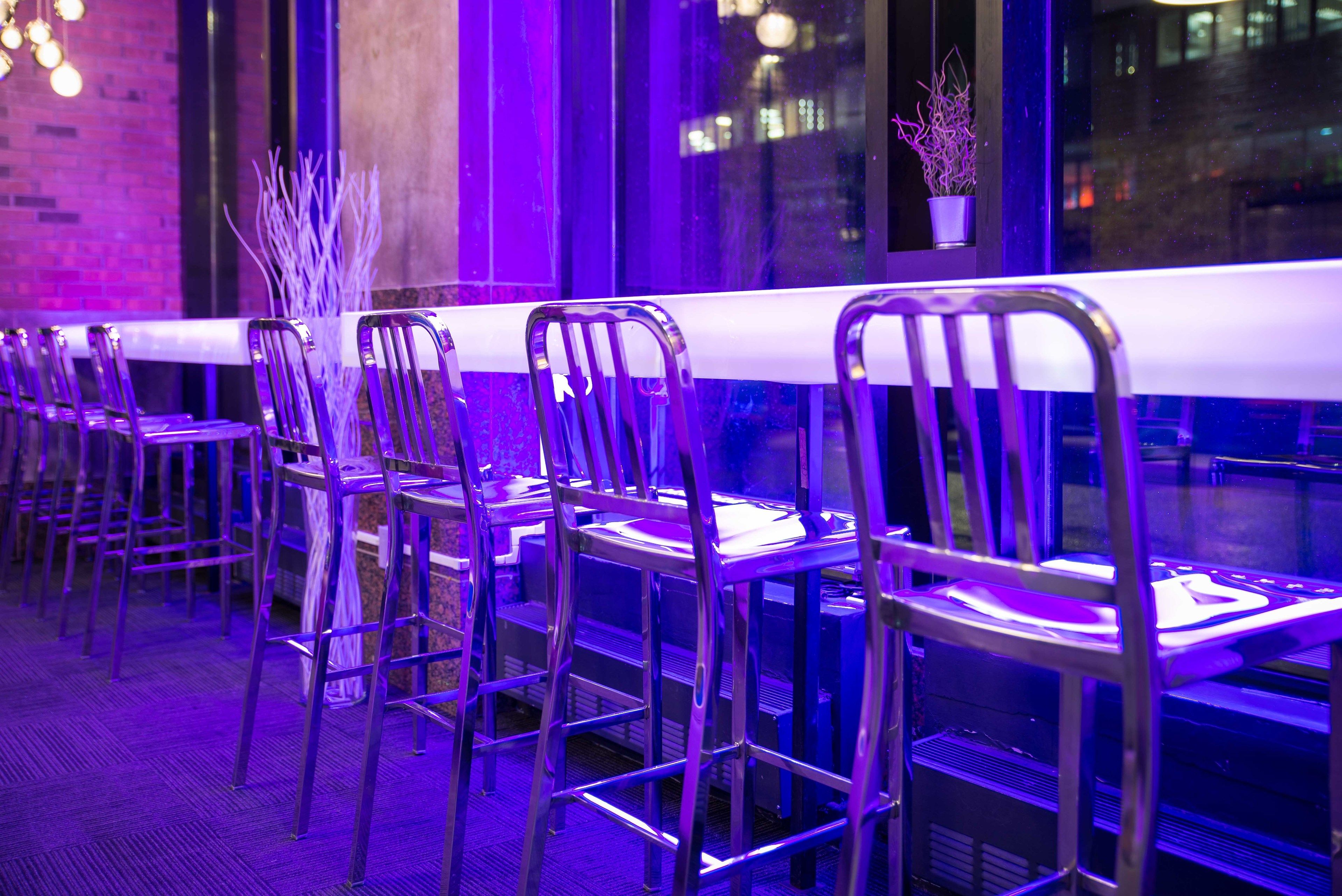 A row of metal chairs with white tablecloths, a potted plant, and a brick wall in the background.
