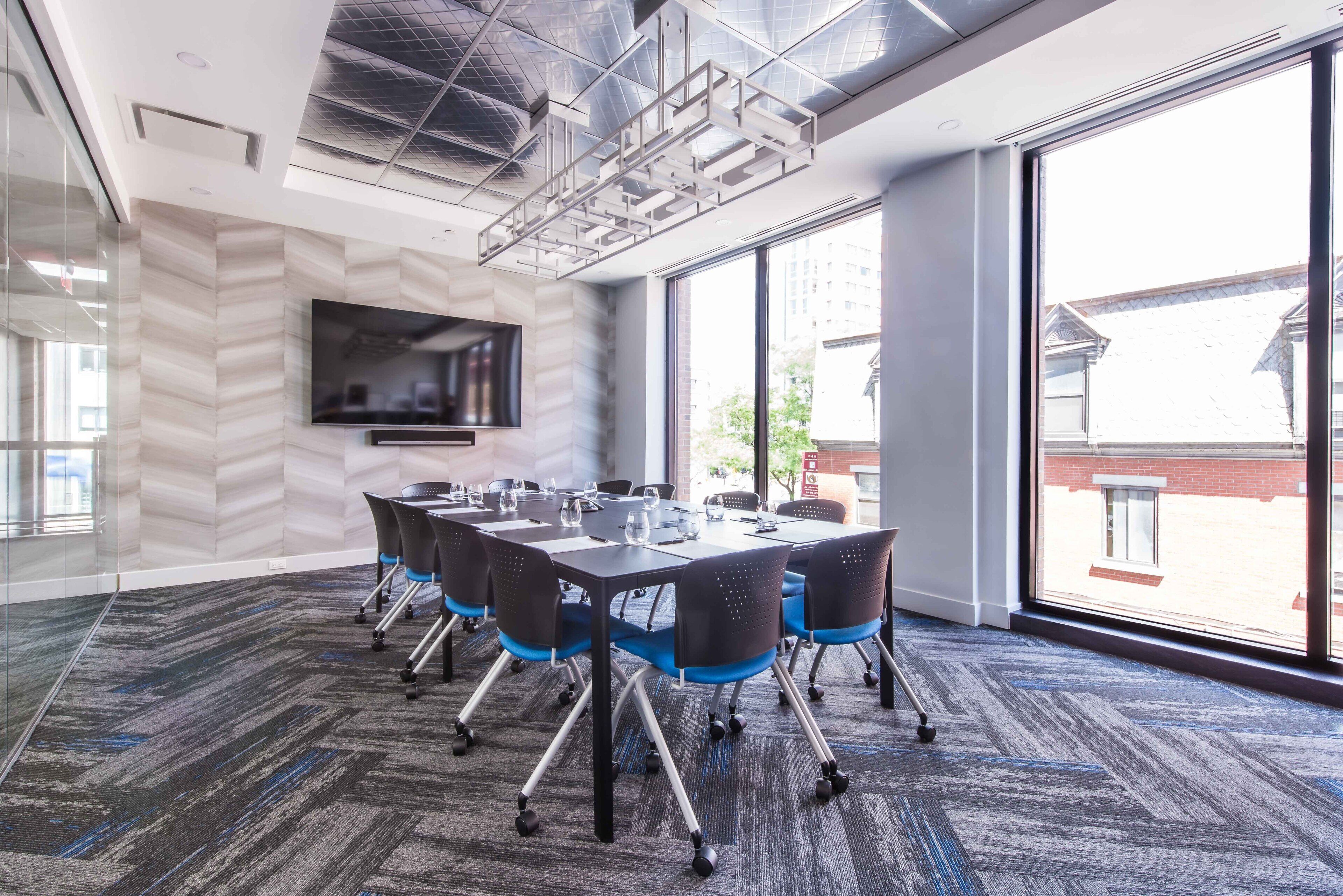 Une salle de conférence équipée d’une grande table, de chaises, d’un téléviseur à écran plat et de grandes fenêtres donnant sur les bâtiments environnants.