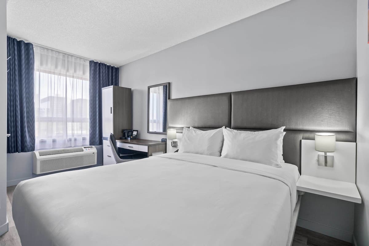 A hotel room with a large bed, bedside tables with lamps, a desk, a mirror, and a window with curtains.