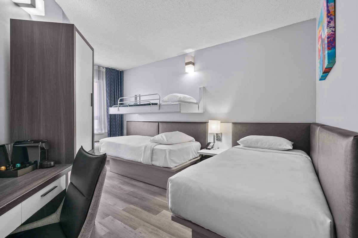 A hotel room with two beds, a desk with a coffee maker, a chair, and a bedside table with a lamp.