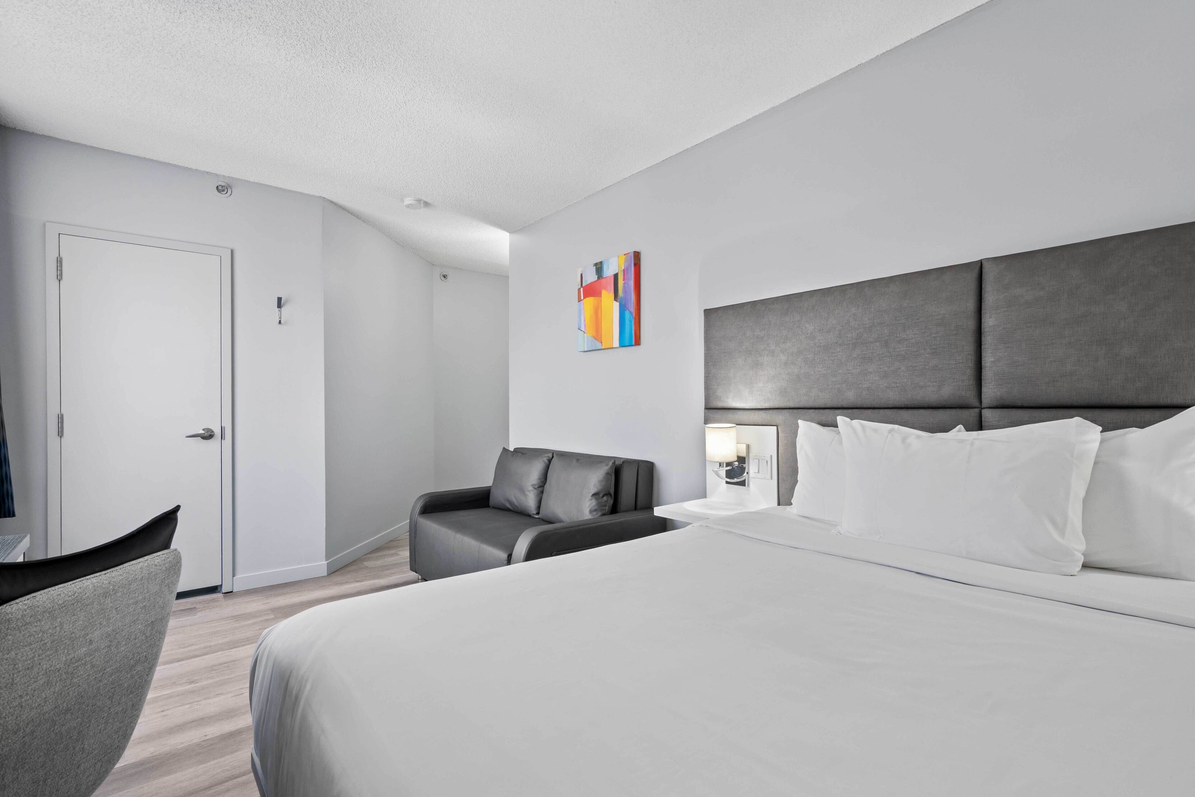 Une chambre d’hôtel moderne avec un grand lit, un canapé gris, un bureau et une porte.