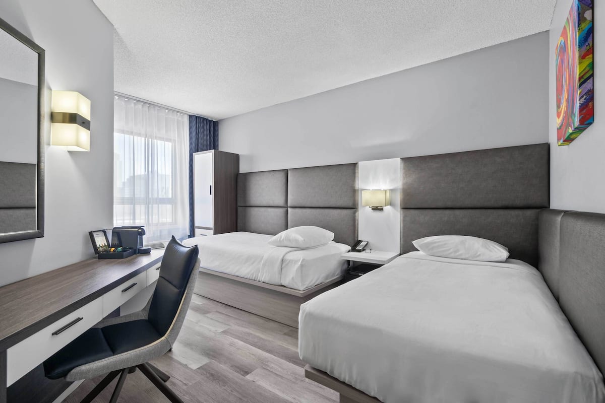 A hotel room with two beds, a desk, a chair, a desk lamp, a phone, and a painting on the wall.