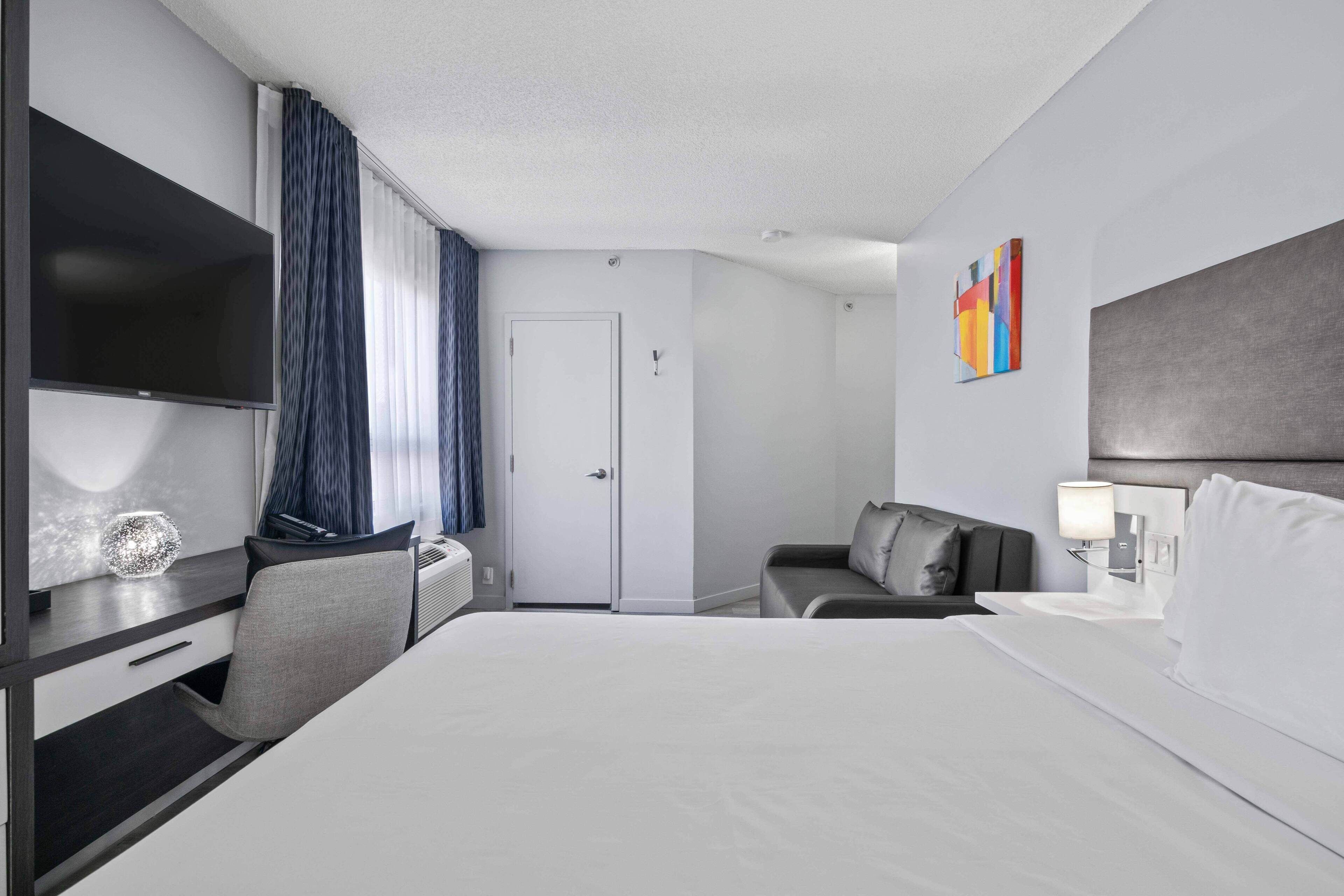 A hotel room with a bed, a desk with a flat-screen TV, a chair, a sofa, a door, and a wall-mounted artwork.