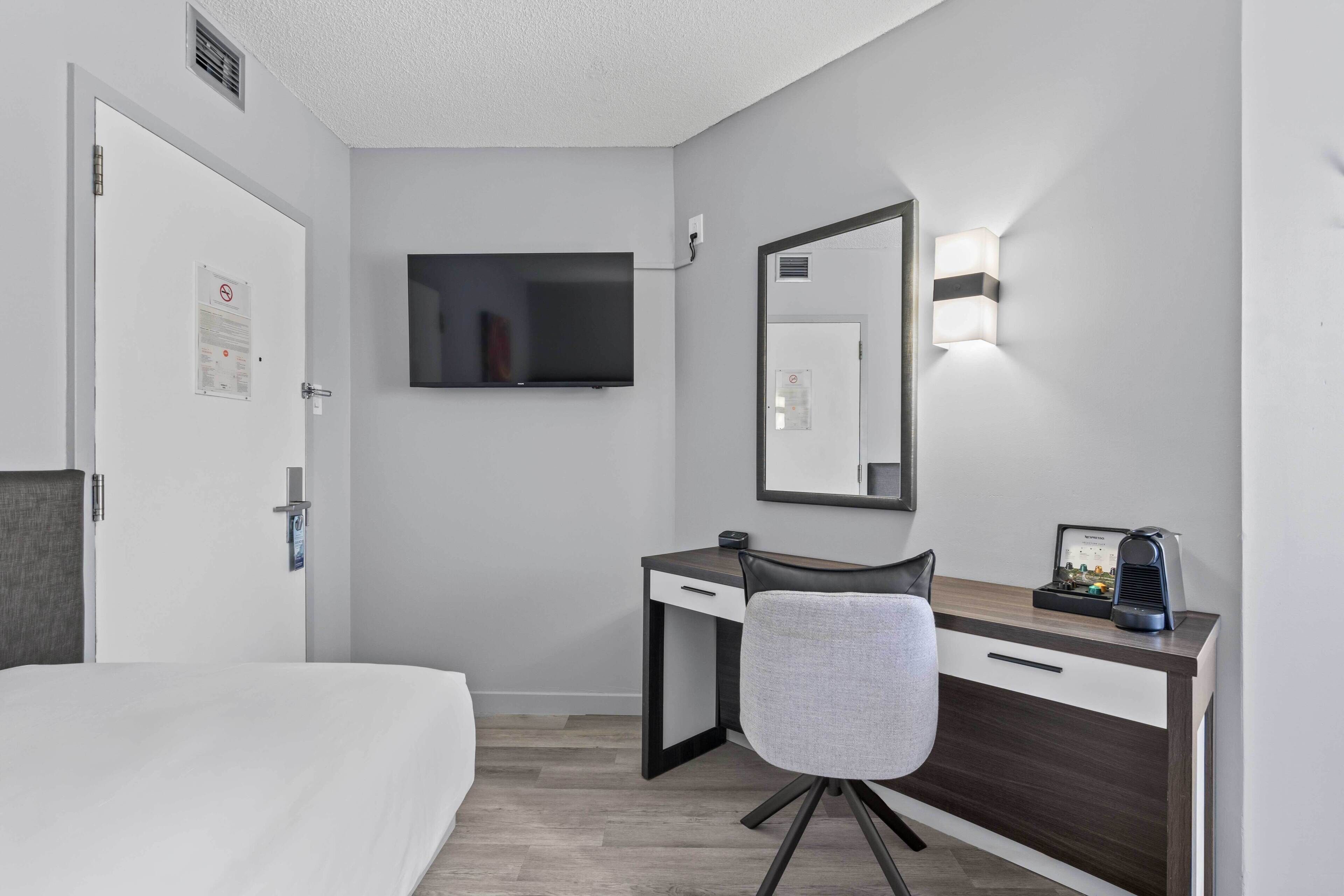 Una habitación de hotel con cama, escritorio con silla, televisor de pantalla plana, espejo y lámpara de pared.