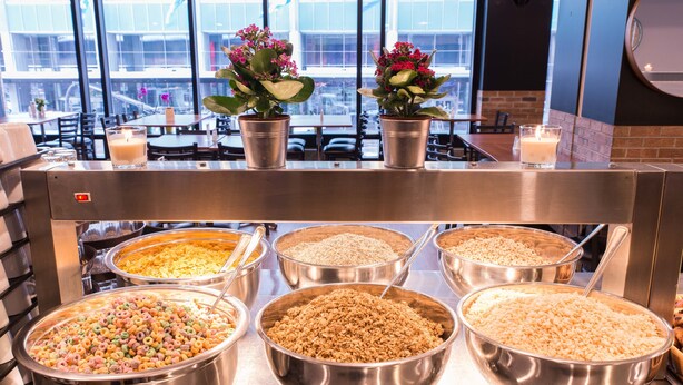 A buffet area with various food options in large metal bowls, potted plants on the counter, and a view of the city through large windows. - Travelodge Hotel by Wyndham Montreal Centre (Montreal)