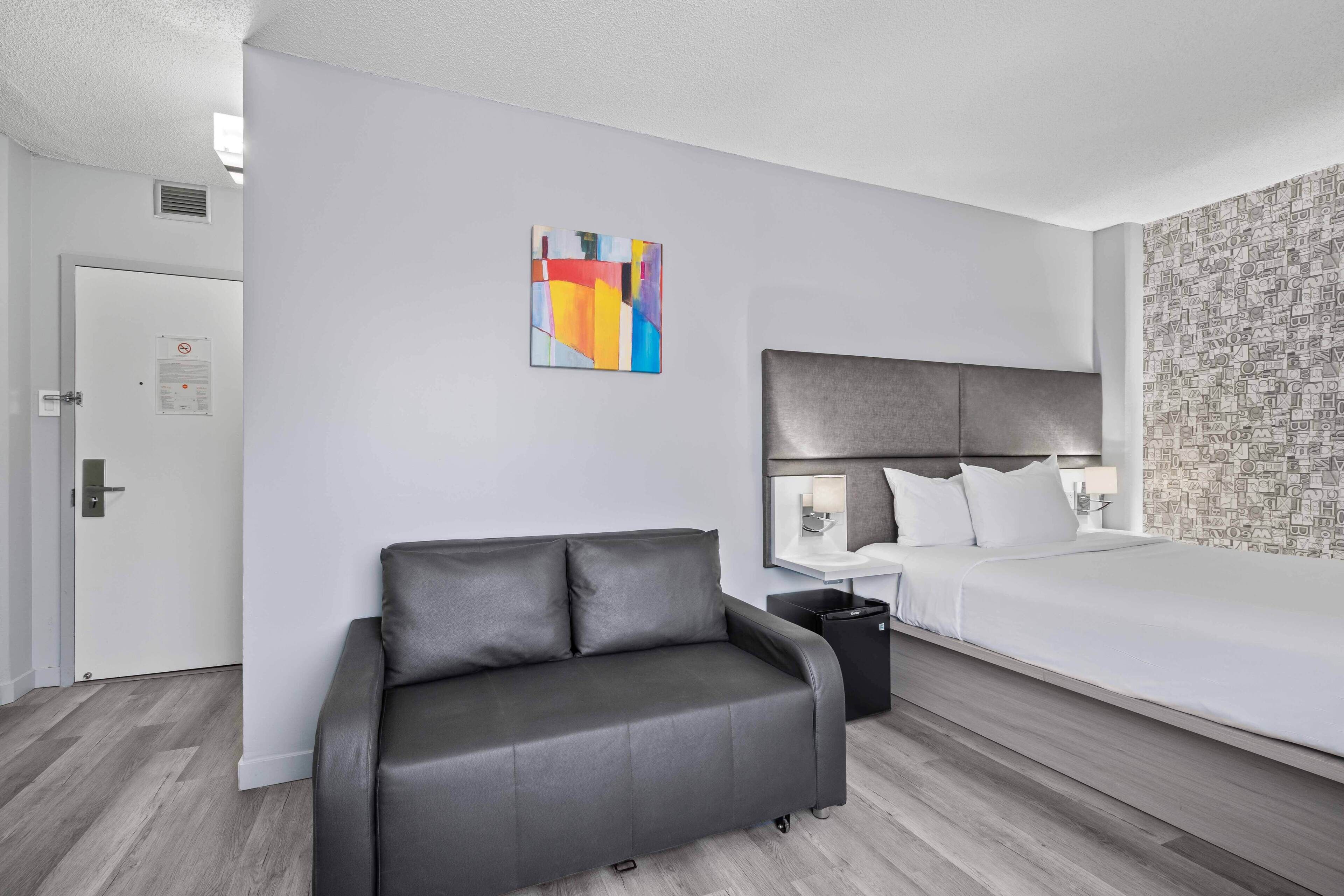 Une chambre d’hôtel moderne avec un canapé gris, un lit avec une literie blanche, une peinture abstraite aux couleurs vives accrochée au mur et un papier peint à motifs sur un des murs.