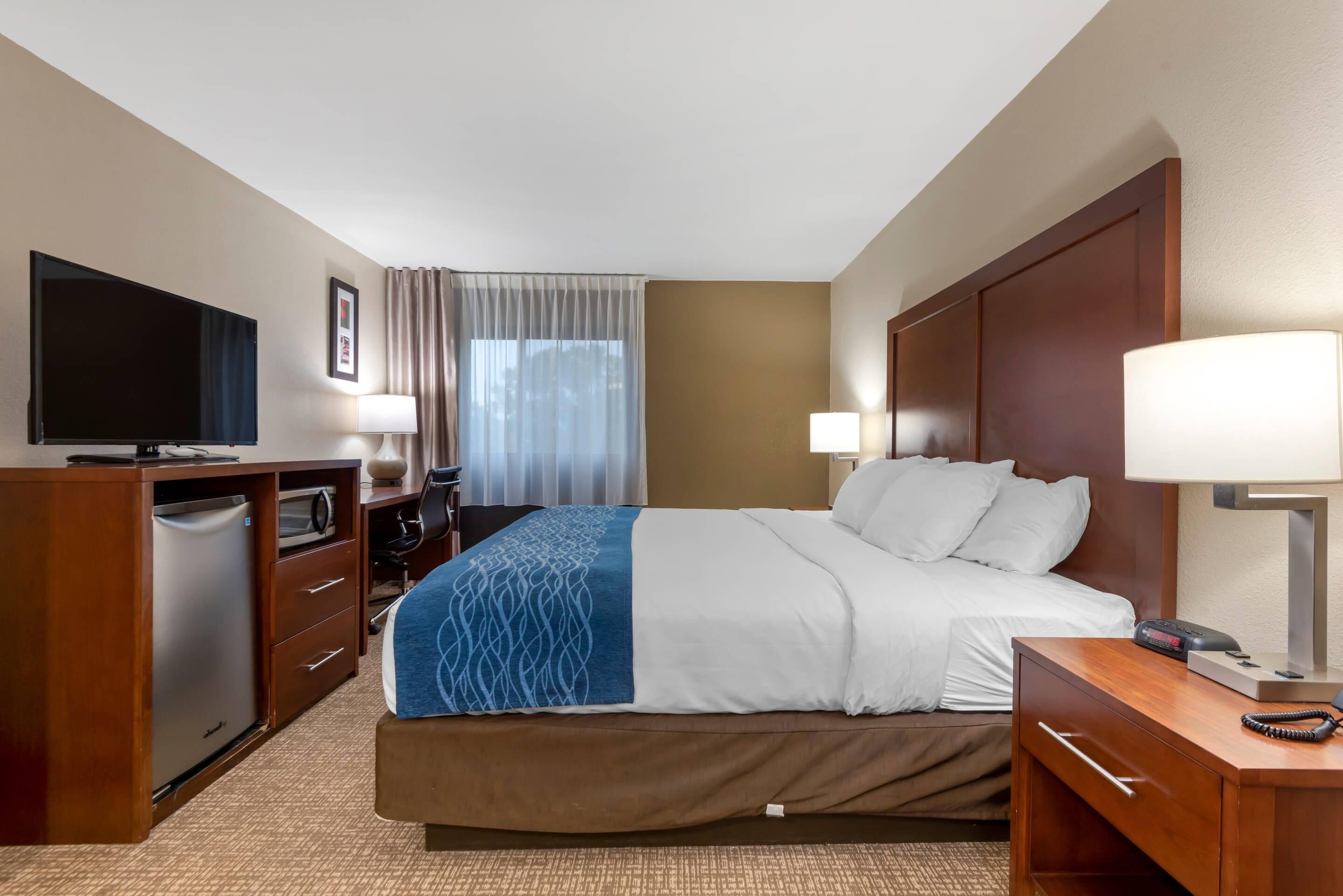 Une chambre d’hôtel comprenant un lit, un bureau avec une télévision, une lampe de bureau, un téléphone et une fenêtre avec des rideaux.
