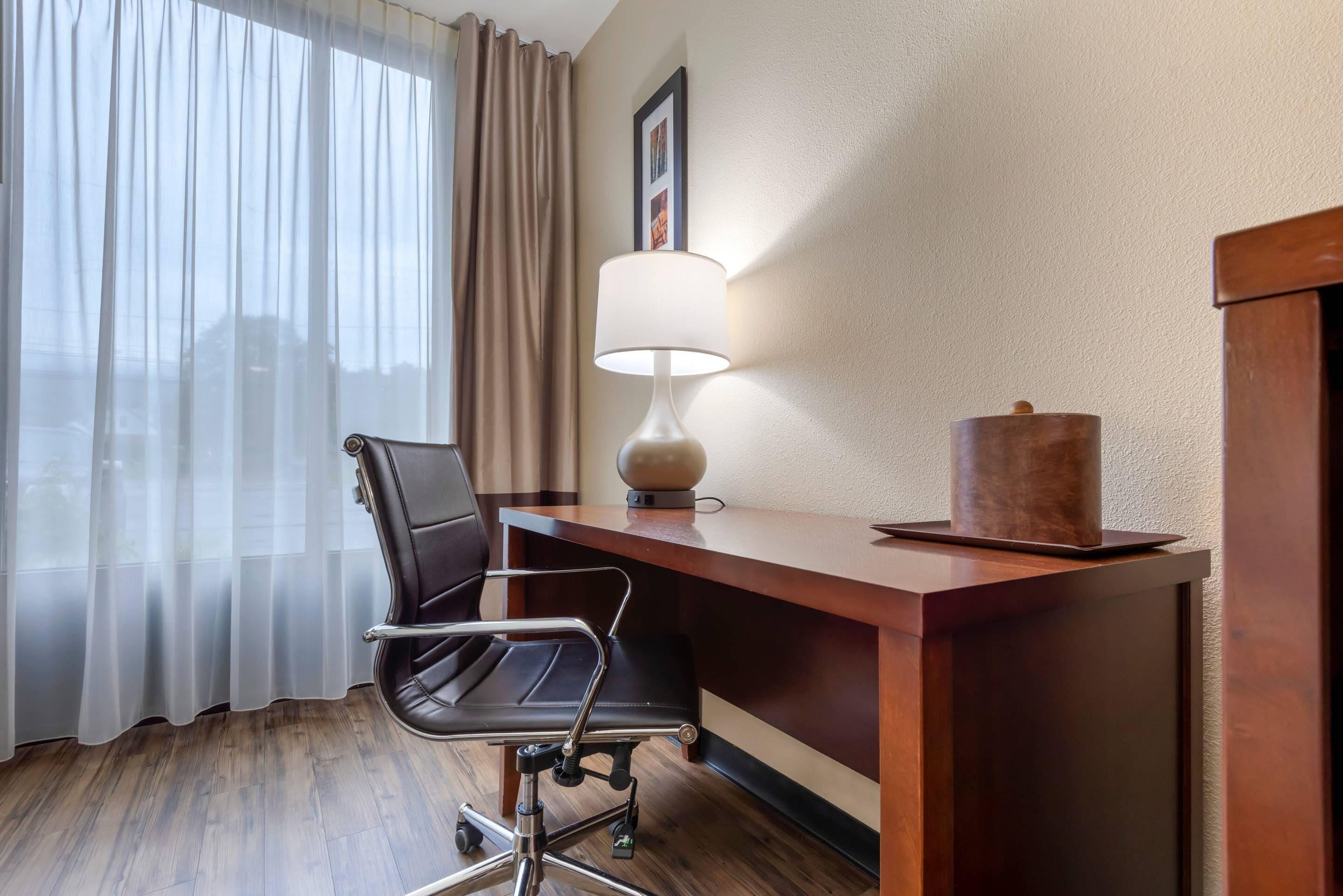 Habitación de hotel con escritorio de madera, una silla de oficina negra, una lámpara y un cuadro en la pared.