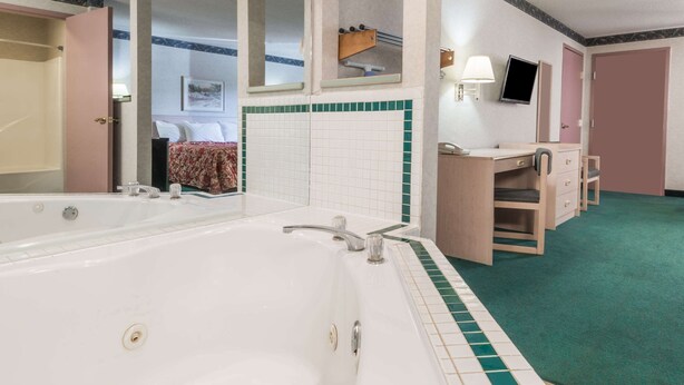 A hotel room with a large bathtub, a vanity with a mirror, a television, and a bed with a floral bedspread. - Knights Inn Ottawa (Ottawa)