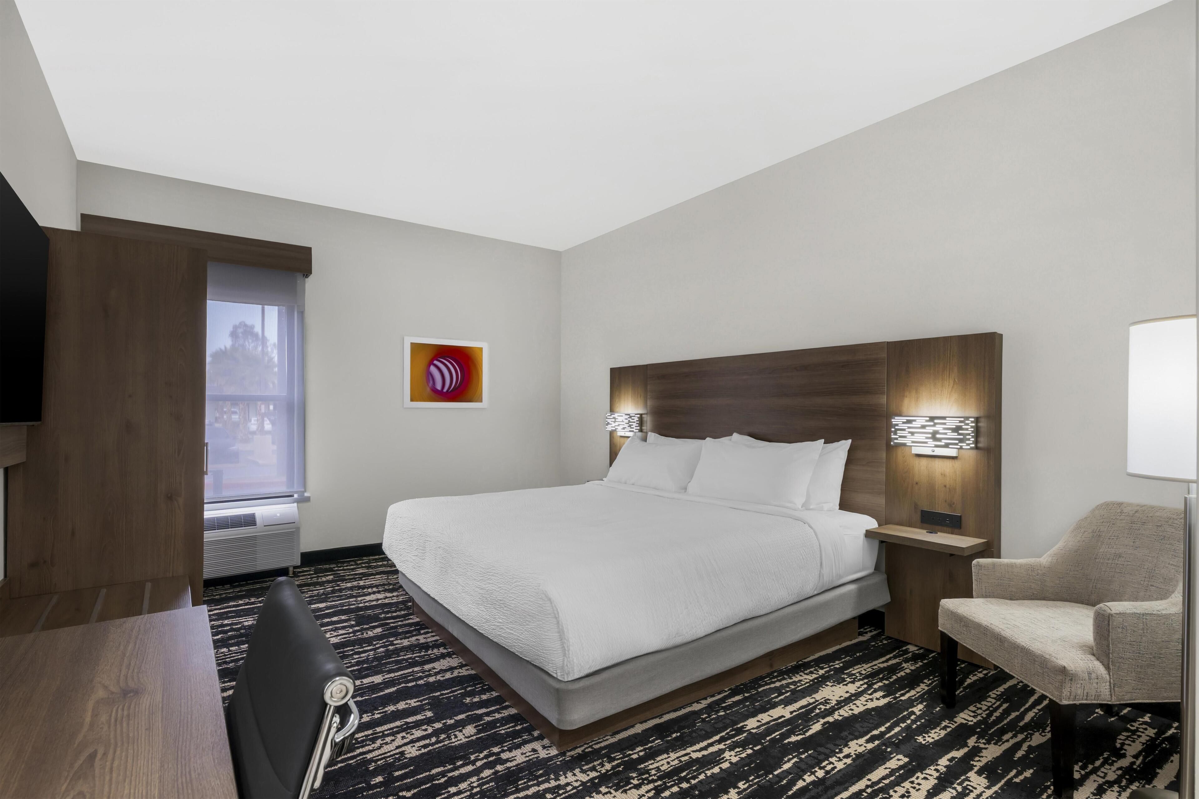 A hotel room with a bed, a chair, a desk, a TV, a window, and a wall art piece.