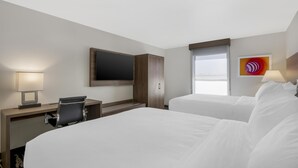 Une chambre d’hôtel avec deux lits, un bureau avec une chaise, un téléviseur fixé au mur et une fenêtre avec des stores.