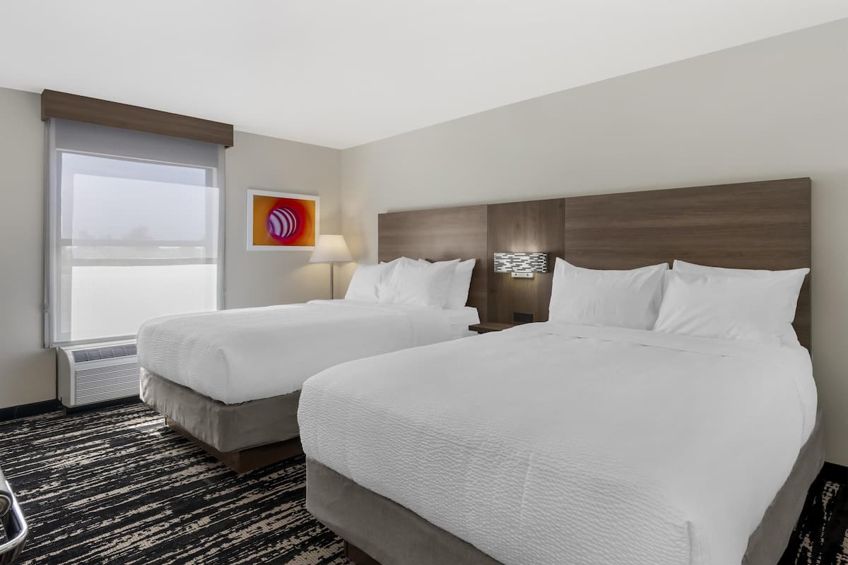 A hotel room with two beds, a window with blinds, a wall-mounted lamp, and a radiator.