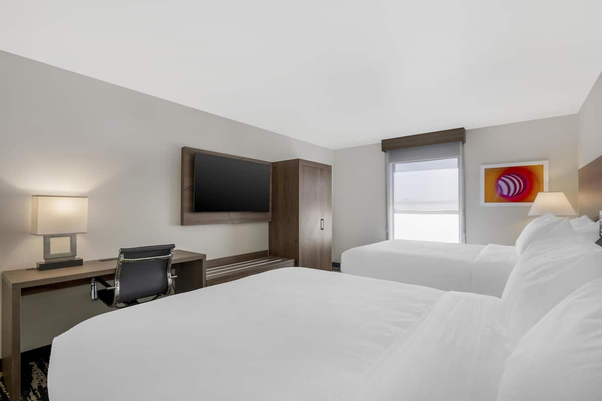 A hotel room with two beds, a desk with a chair, a flat-screen TV mounted on the wall, and a window with a view.