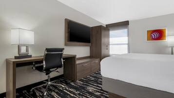 A hotel room with a bed, desk, chair, TV, and artwork on the wall.