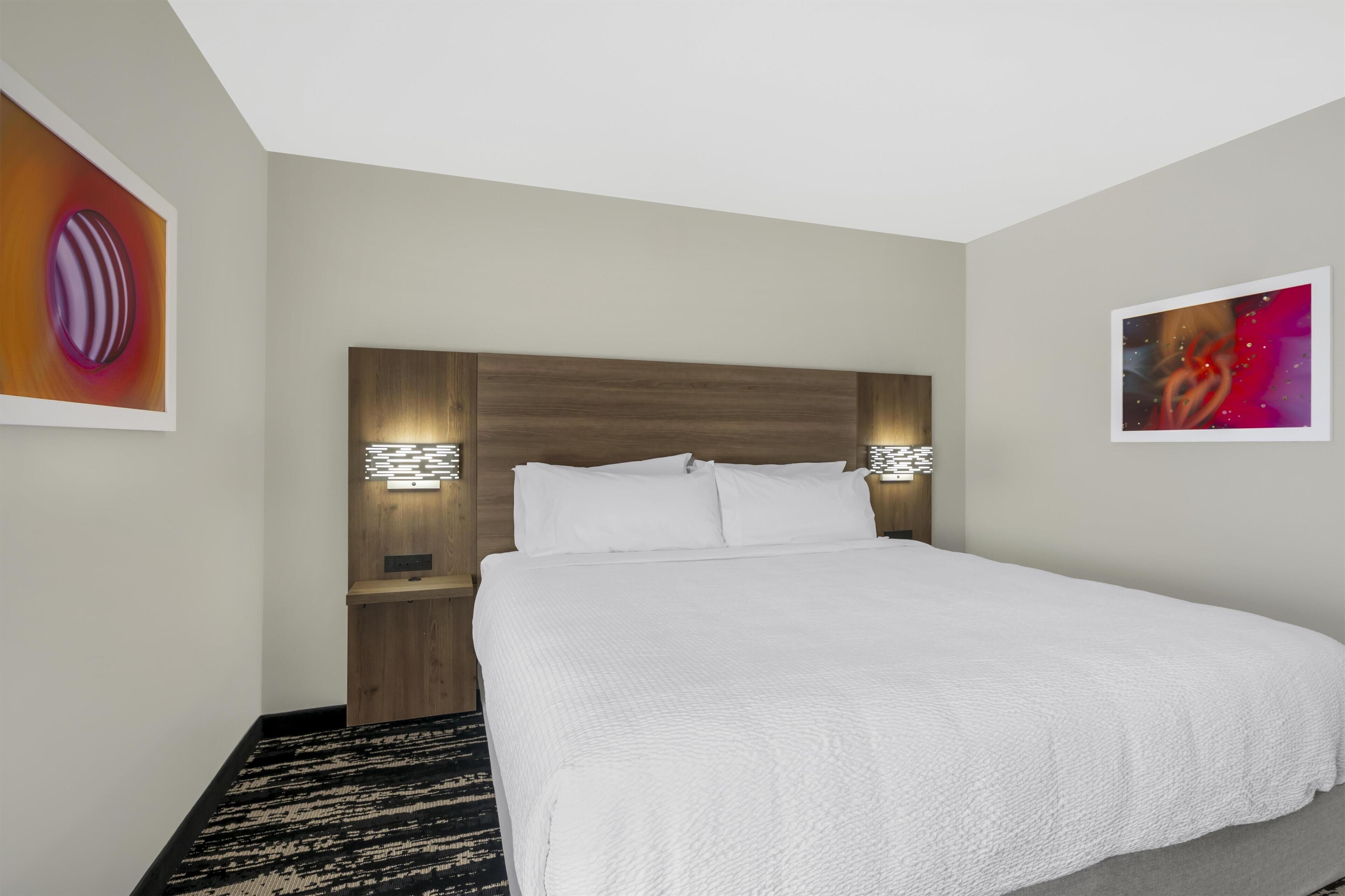 A hotel room with a large bed, a wooden headboard, two bedside tables with lamps, and abstract wall art.