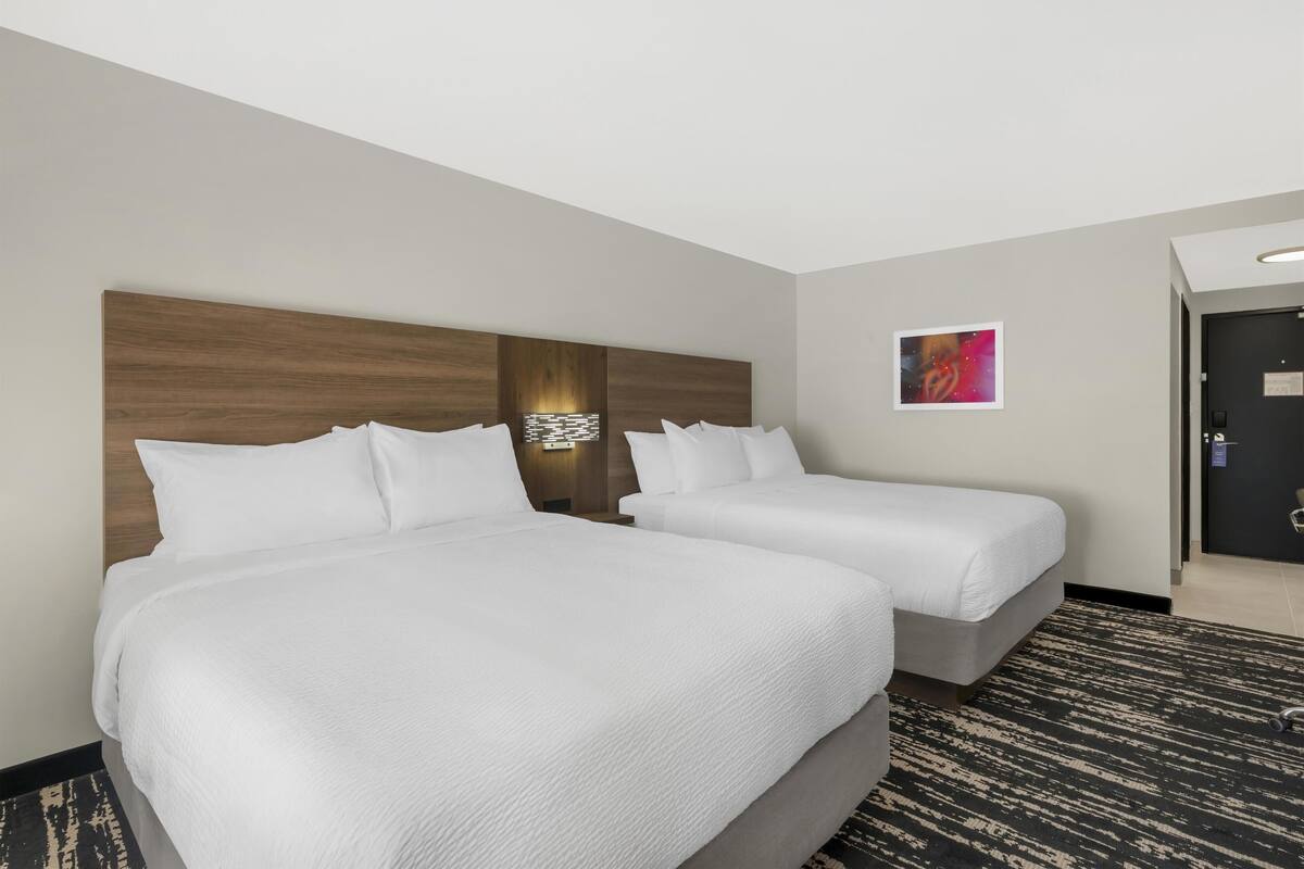 A hotel room with two beds, a headboard with a light fixture, a wall-mounted artwork, and a carpeted floor.