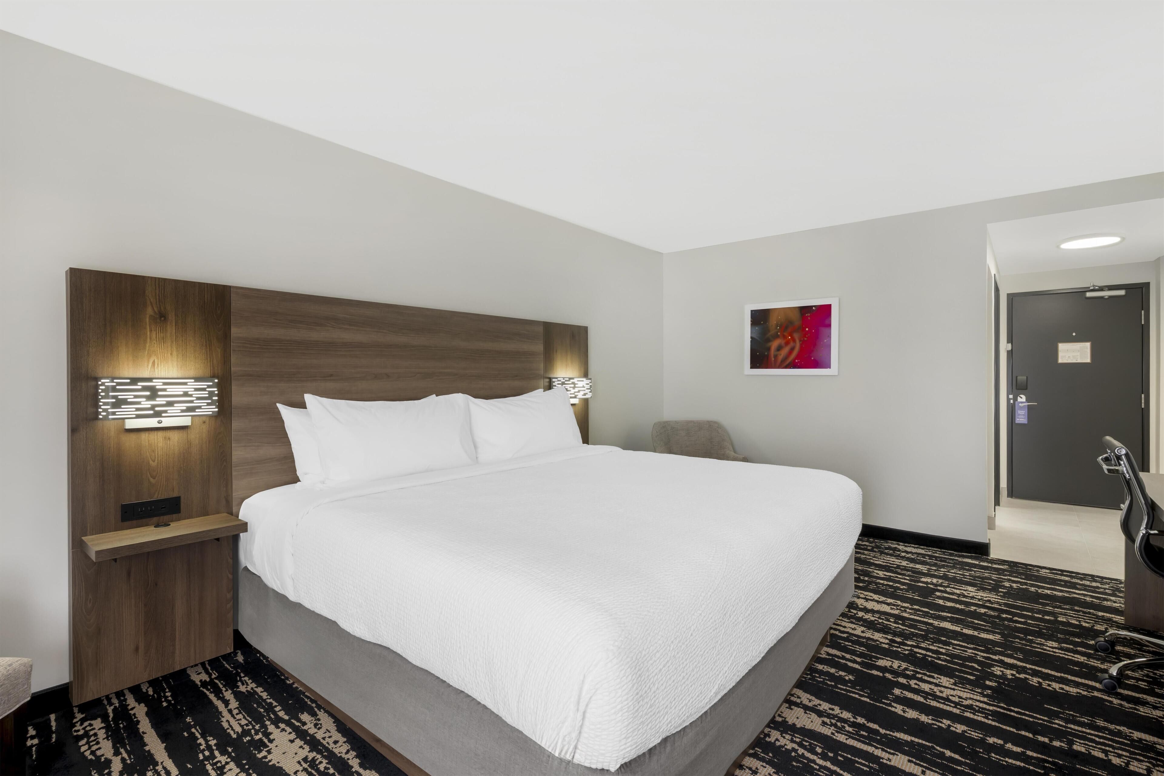 A hotel room with a large bed, a wooden headboard, bedside lamps, a small table, a chair, and a carpeted floor.