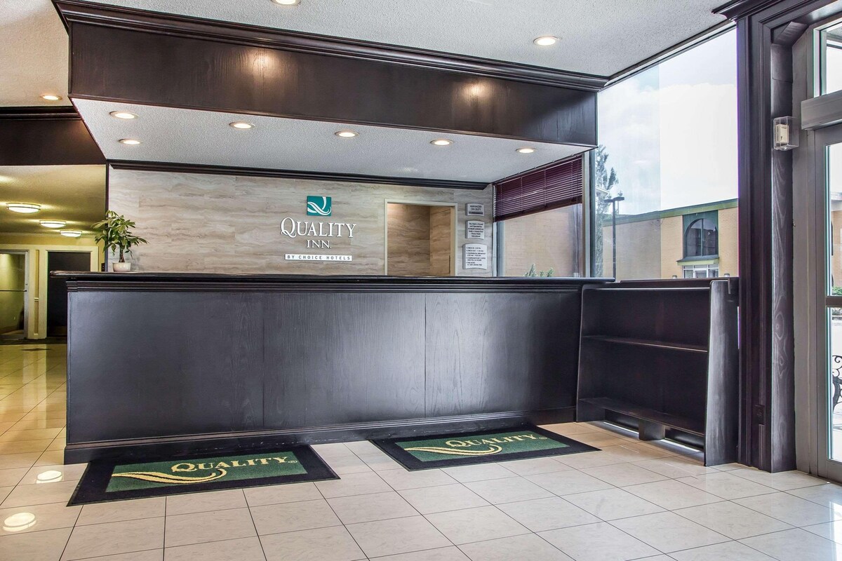 A hotel reception desk with a marble top, a sign reading 'Quality Inn by Choice Hotels,' and a 'Manager on Duty' sign. There are two doormats with the hotel logo on the floor.