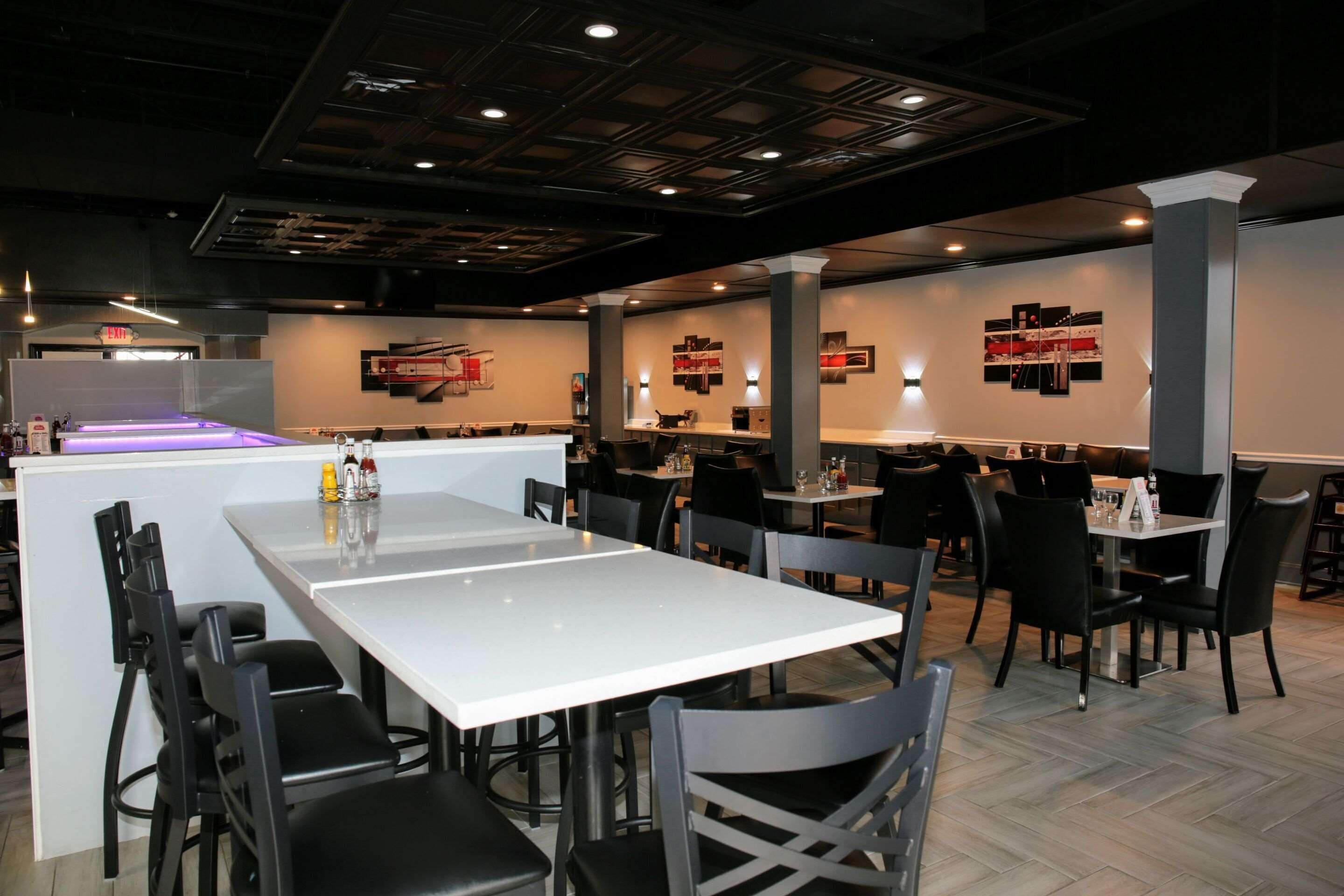 A restaurant with white tables and black chairs, a bar area with a purple light, and artwork on the walls.